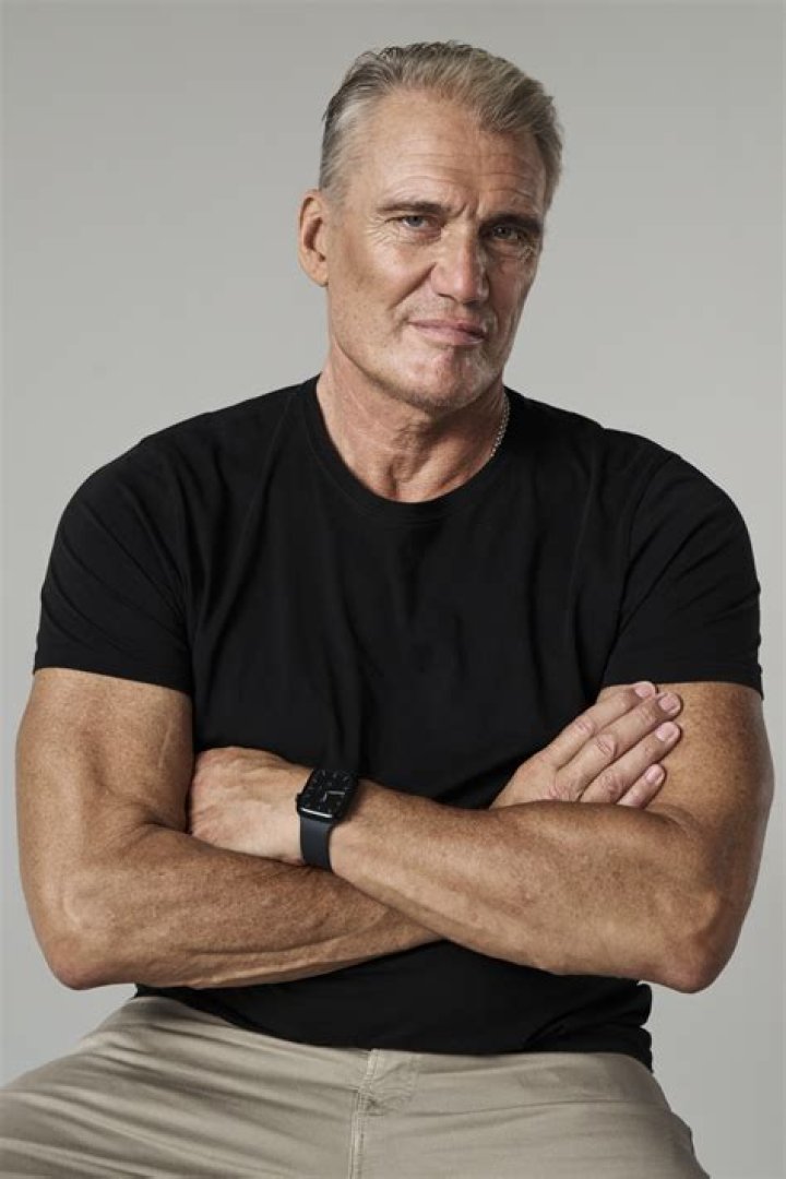 Deepfake Dolph Lungren with an '80s flat top wearing a white tank top clutches a metal railing and reaches down.